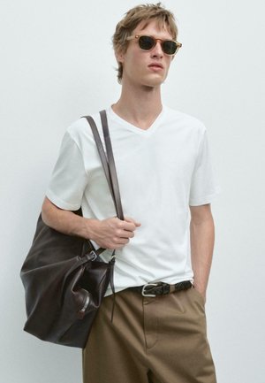 Jonge man draagt beige broek en wit T-shirt met V-hals, met een grote donkerbruine leren tas over zijn schouder, en draagt een zonnebril.