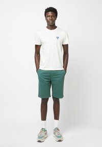 T-Shirt blanche à manches courtes avec un graphisme en forme de cœur, shorts verts et baskets. Le modèle se tient avec les mains dans les poches devant un fond uni.