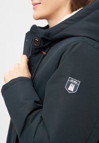 Marinblå jacka med en fuskpälsfoder huva, bruna knappdetaljer och en sköldpatch med en torndesign på vänster ärm.