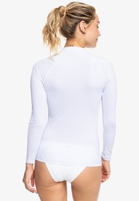 White long-sleeve rash guard, smooth texture, fitted design. Features flat seams, round neckline, and logo detail on upper back.