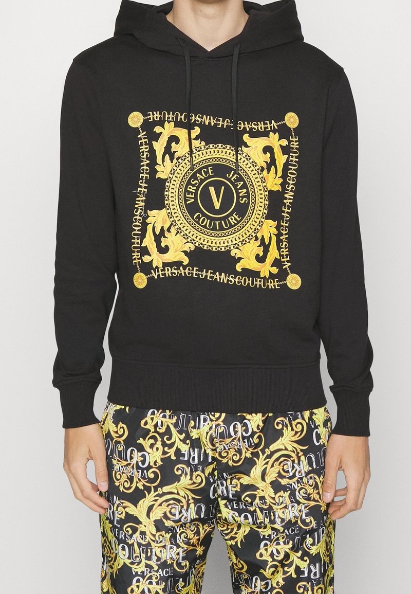 Černá mikina s kapucí s centrálním zlatým motivem s propracovanými vzory a textem, doplněná černými a zlatými vzorovanými kalhotami.