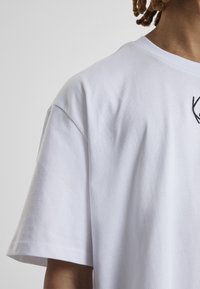 T-shirt en coton blanc à manches courtes, comportant un simple motif noir brodé sur le haut à gauche de la poitrine, texture lisse.