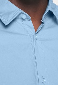 Ljusblå skjorta med krage, som har synliga sömmar, knappar och en mjuk tygtextur.