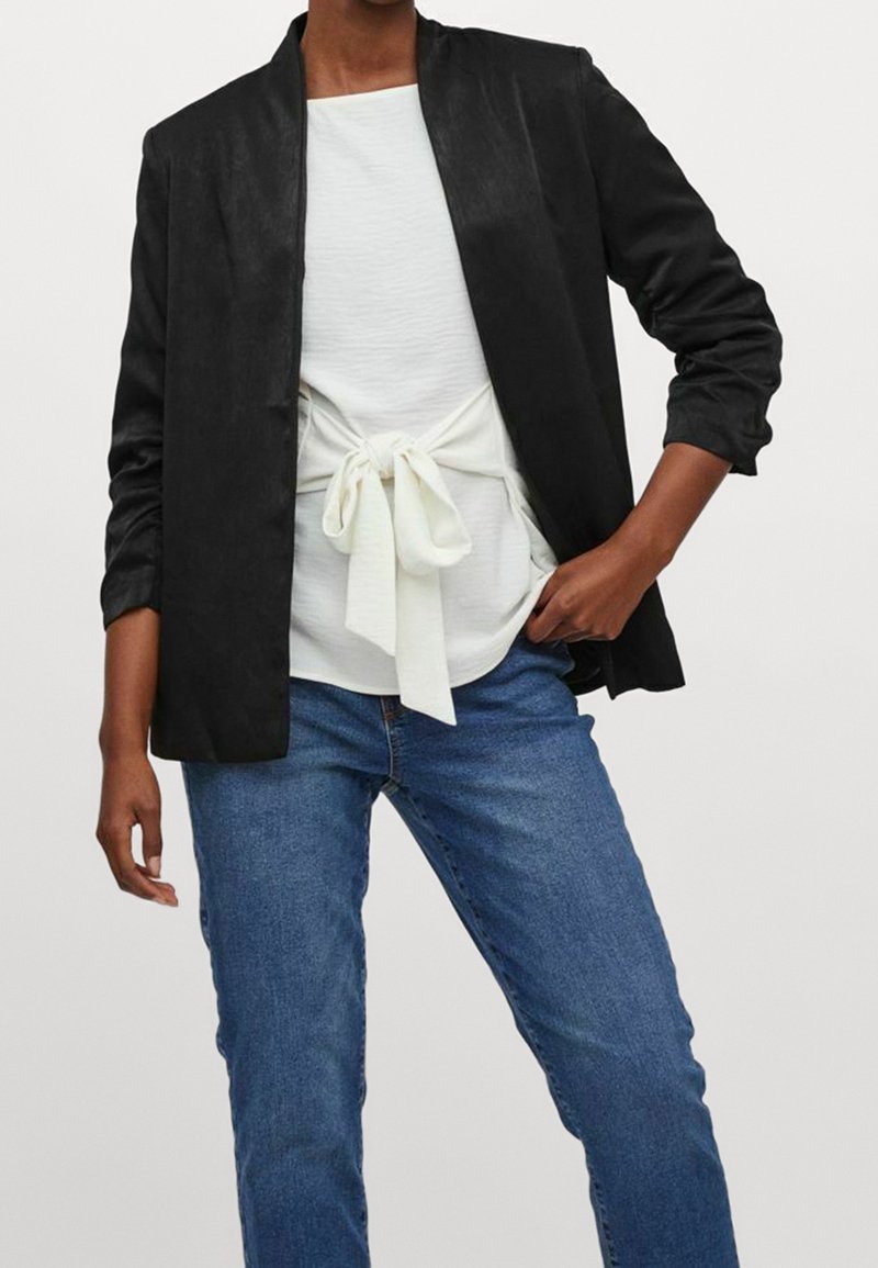 Personne portant un blazer noir ouvert, un haut blanc avec un nœud à l'avant et un jean bleu, debout devant un fond clair uni.