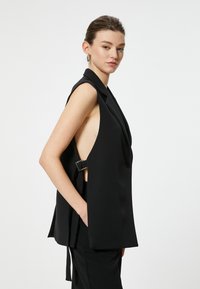 Blazer senza maniche nero con un profondo taglio laterale, rever strutturati, bottone frontale lucido e dettaglio con cintura regolabile in vita.