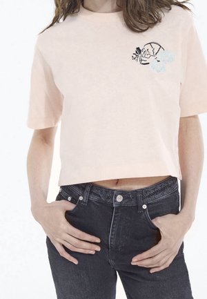 Persona que lleva una camiseta corta de color rosa claro con un pequeño diseño de calavera y flor en blanco y negro en el pecho, combinada con vaqueros oscuros de cintura alta.