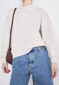 Jasnoszara bluza z okrągłym dekoltem i logo na plecach, zestawiona z wysokim stanem niebieskich jeansów oraz małą brązową torebką na ramię.