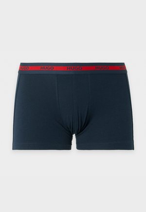 Sportlicher dunkelhäutiger Mann steht barfuß, trägt marineblaue Boxershorts mit orangefarbenem Bund, Hände hinter dem Rücken, blickt seitlich auf weißem Hintergrund.
