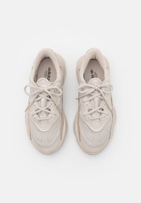 Ljusbeiga träningsskor med ovandel i mesh, rund tå och dämpad sula. Har snörning och texturerade paneler på sidorna.