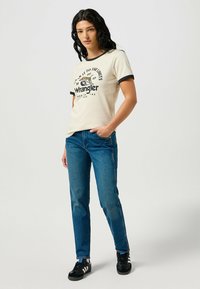 Camiseta crema con ribetes negros y estampado gráfico, combinada con jeans azul y zapatillas negras. Diseño simple, ajuste casual y textura textil evidente.