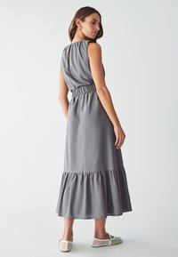 Vestido maxi de algodón gris con cintura fruncida, diseño sin mangas, cuerpo plisado y dobladillo en capas con volantes. Combinado con zapatos de malla blanca.