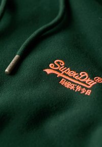 Felpa con cappuccio verde scuro in tessuto morbido, con logo "SuperDry" ricamato in arancione e una placchetta metallica sui lacci.