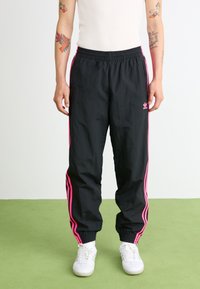 Pantaloni sportivi neri con strisce rosa sui lati, vita elasticizzata e caviglie a coste. Presentano il logo adidas sulla coscia sinistra. Tessuto dalla texture liscia.