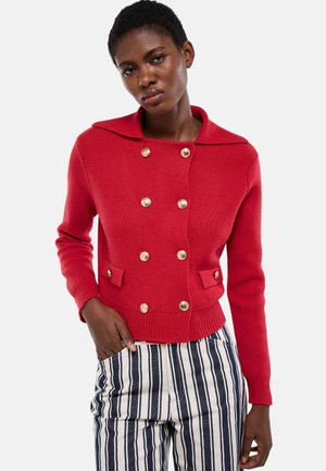 Femme portant un cardigan rouge à double boutonnage avec des boutons dorés et un pantalon blanc à rayures verticales noires, debout avec une main dans la poche.
