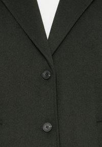 Mørkegrønn ullblandingsblazer med spiss krage og to sorte knapper foran, med en myk tekstur og strukturert silhuett.