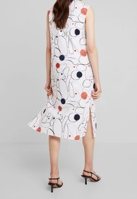 Vestito senza maniche in tessuto bianco, caratterizzato da pattern astratti in nero, rosso e blu navy. Il dettaglio con spacco laterale accentua il movimento. Tacchi neri con cinturini.