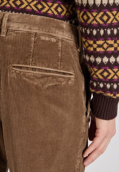 Brązowe spodnie z korduru o subtelnej fakturze, z tylną kieszenią i haftowanym logo marki. W komplecie z dzianinowym swetrem w desenie.