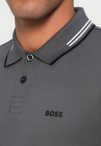 Szara koszulka polo z teksturowanej tkaniny, z czarno-białym paskowanym kołnierzykiem, czarnymi guzikami oraz czarnym logo "BOSS" na piersi.