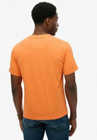 Camiseta de algodón naranja con cuello redondo, mangas cortas y una textura suave, vista desde la parte de atrás sobre un fondo liso.