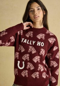 Junge Frau, die einen kastanienbraunen Pullover mit rosa Pferdeköpfen, dem weißen Text "TALLY HO" und einem weißen Hufeisen trägt und vor einem neutralen Hintergrund posiert.