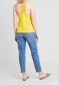 Gul linne med spetsdetalj på baksidan, matchat med ljusblå jeans och mönstrade sandaler. Har en figursydd design och justerbara axelremmar.