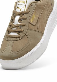 Beige mockasin sneakers med vit gummisula, försedd med guld Puma-logotyp, platta snörning och texturerade sidodetaljer. Lätt förlängd form.