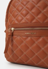 Mochila acolchada marrón hecha de cuero sintético texturizado. Presenta herrajes dorados y un bolsillo frontal con cremallera con el logo "ANNA FIELD".