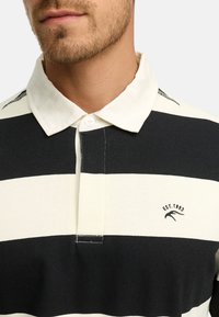 Polo a righe nero e crema con colletto alla francese, dotato di un dettaglio logo ricamato "EST. 1993" sul lato sinistro.