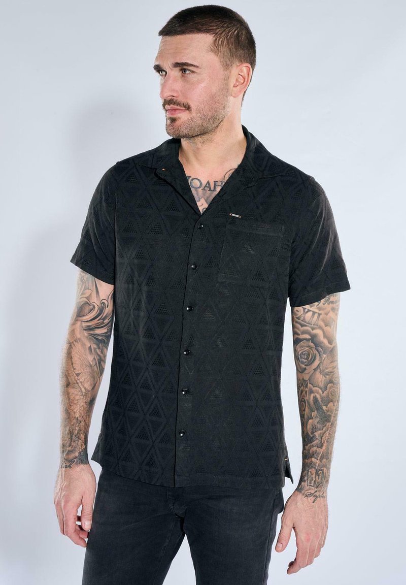 Man with short hair and tattoos on both arms wearing a black short-sleeve button-up shirt with geometric patterns.