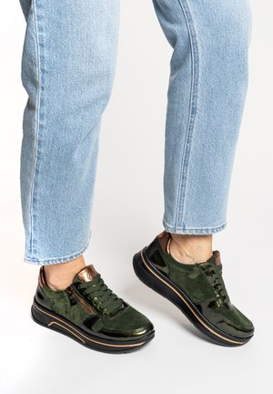 Personne portant un jean bleu clair et des baskets vert foncé avec des semelles noires et des accents bronze, debout sur une surface blanche.