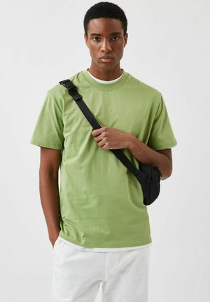 Grünes Kurzarm-T-Shirt mit rundem Ausschnitt, kombiniert mit weißen Hosen und einer schwarzen Umhängetasche mit Reißverschlussdetails.