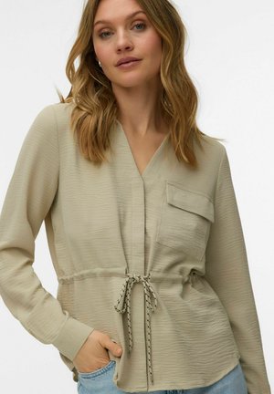 Femme aux cheveux blonds mi-longs portant un chemisier beige texturé avec une poche avant et une taille à cordon, jean bleu clair.