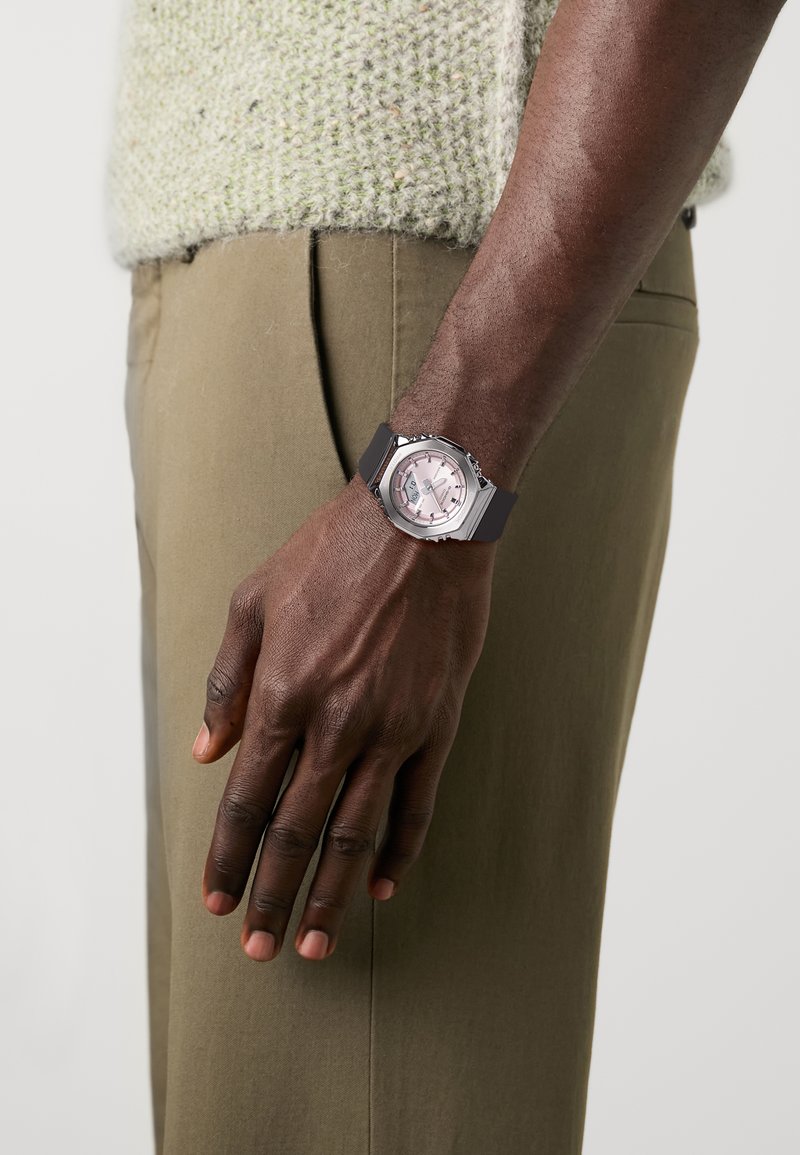 Montre analogique avec un boîtier en métal argenté, un cadran rose et un bracelet en silicone noir. Portée sur une main avec un teint de peau texturé et sombre.