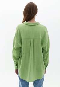 Zielona, teksturowana koszula z kołnierzykiem, guzicznymi mankietami i zakładką z tyłu. Materiał wydaje się lekki i nieco przezroczysty.