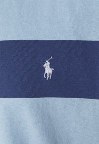 Pikétröja med horisontella ränder i ljusblått och marinblått, tillverkad av bomull med en mjuk textur. Broderad vit logotyp av en häst och ryttare.