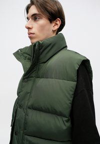 Roheline puffermantel kõrge kraega, nööbiga kinni keeratav ja sileda tekstuuriga, kantud musta pika varrukaga särk üle, külgvaatest.