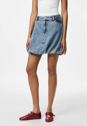 Jupe en denim bleu clair avec une taille ajustée, fermeture à bouton et ourlet froncé. Associée à des ballerines rouges à détails de sangle.