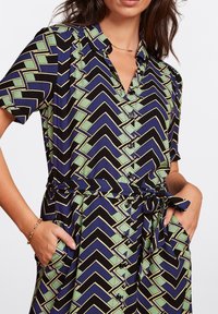 Short-sleeve dress in navy with green and black zigzag pattern. Features a button-down front and a tied waist with pockets. Smooth fabric.