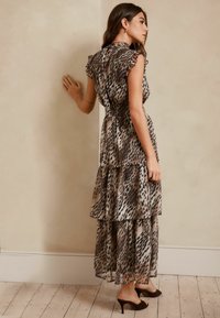 Slojna haljina od lagane, prozirne tkanine s uzorkom leopard printa u bež i smeđim tonovima. Ruffles oko vrata i pripijeni struk, s potpeticama vidljivim.
