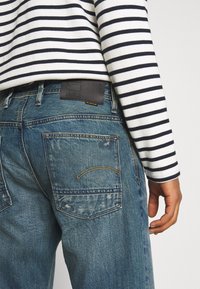 Wyblakłe niebieskie dżinsy o prostym kroju, z tylnymi kieszeniami, ciemną skórzaną naszywką marki oraz paskowaną bluzką z długim rękawem.