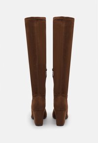 Botas marrones de ante por encima de la rodilla con tacón cuadrado, que cuentan con costuras verticales y un cierre de cremallera lateral.