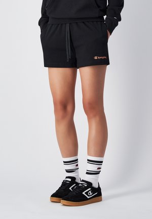 Pantalones cortos atléticos negros con cordón, con un logo de Champion en naranja. Combinados con zapatillas negras y calcetines blancos a rayas.
