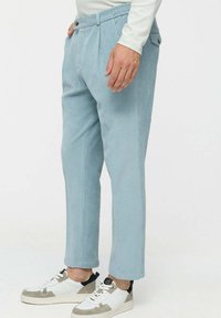 Helle blaue Cordhosen mit Falten, ausgestattet mit einem Bund mit Knopfverschluss und Seitentaschen. Getragen mit weißen und grauen Turnschuhen.