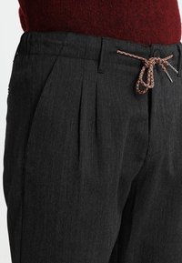 Primo piano di pantaloni grigio scuro con texture e pieghe, con un laccio arancione e nero legato a fiocco, abbinato a un maglione a maglia rossa.