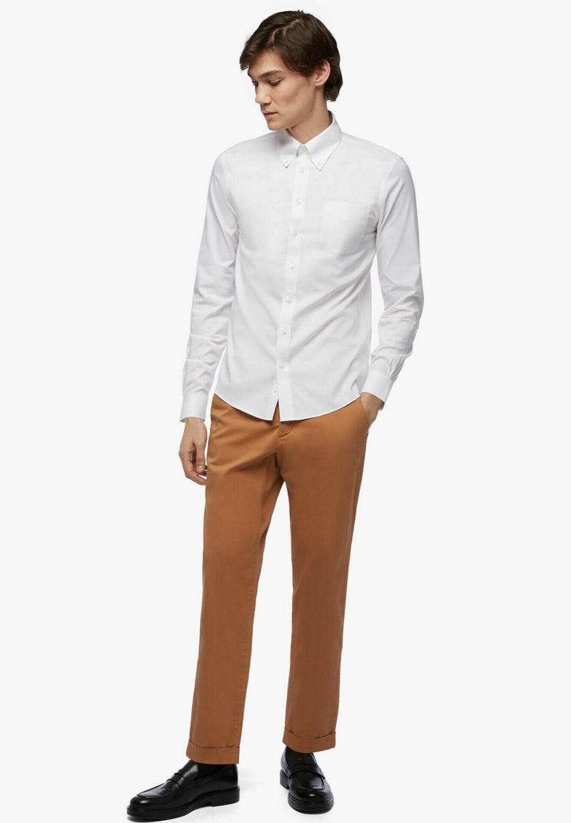 Jonge man in een wit overhemd met knopen, bruine broek en zwarte instappers, staande met één hand in zijn zak, kijkend naar beneden naar de zijkant.