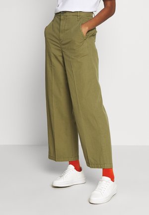 Person trägt olivfarbene, weit geschnittene Hose, weiße Turnschuhe und leuchtend orangefarbene Socken, steht mit den Händen in den Taschen vor einem schlichten Hintergrund.