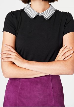 Persona con los brazos cruzados, vestida con una blusa negra con cuello blanco a rayas y una falda morada, vista desde los hombros hasta la cintura.