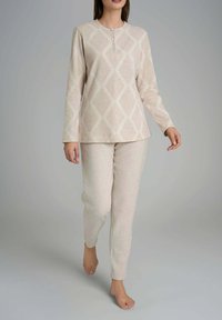 Donna che cammina a piedi nudi indossando un top beige a maniche lunghe con motivo a diamante e pantaloni coordinati su uno sfondo grigio.