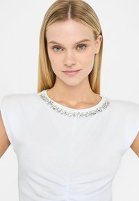 T-shirt bianco a maniche corte, con una texture arricciata e un colletto impreziosito da cristalli trasparenti.