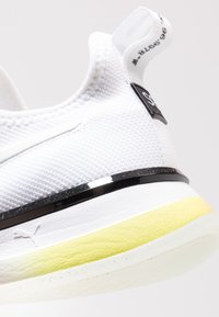 Białe sneakersy o teksturowanej powierzchni z czarnym paskiem akcentowym, żółtym detalem na pięcie i pętlą do zakupu. Charakteryzują się logo PUMA w pobliżu podeszwy.